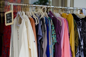 A clothing rack filled with various colorful garments, including dresses and shirts on hangers. A small blackboard sign indicates a sale with prices and discounts.