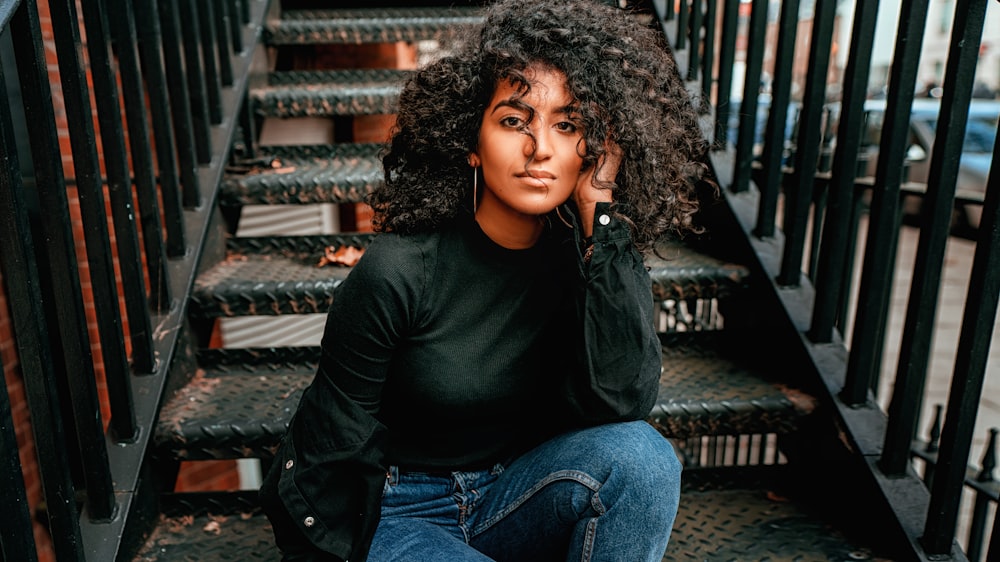 woman in black long sleeve shirt and blue denim jeans sitting on black wooden stairs