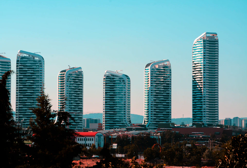 The image presents a cityscape with six modern, tall buildings painted in shades of white and blue, standing against a clear sky. The tallest building is on the left side of the frame, while the shortest one is on the right. In the background, there are trees that add a touch of nature to the urban setting.