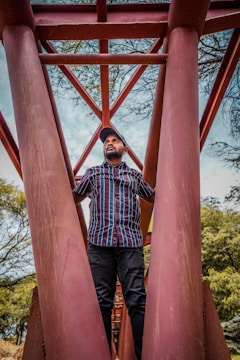 A person is standing under a large red metal framework that resembles the understructure of a bridge. The individual is wearing a striped shirt and a cap, with their hands holding onto the metal beams. Surrounding the framework, there are green trees and a blue sky visible in the background.