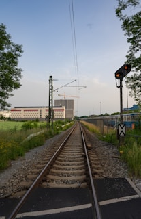 Railway tracks extend into the distance, flanked by greenery on one side and a building with a crane in the background. Overhead power lines run parallel to the tracks. A signal light is visible to the right, with a clear sky above.