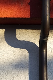 A textured wall with a red surface at the top and a light-colored wall beneath it. A black drainpipe runs vertically along the wall, creating a curved shadow in the sunlight.