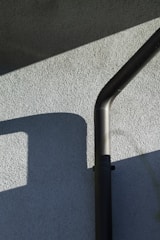 An exterior wall with a textured surface is partially lit by sunlight, casting geometric shadows. A black downspout runs vertically, bending at the top.