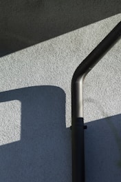 An exterior wall with a textured surface is partially lit by sunlight, casting geometric shadows. A black downspout runs vertically, bending at the top.