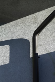 An exterior wall with a textured surface is partially lit by sunlight, casting geometric shadows. A black downspout runs vertically, bending at the top.