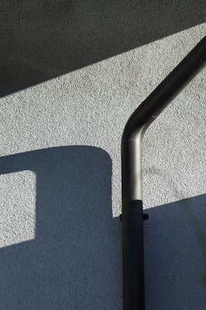 An exterior wall with a textured surface is partially lit by sunlight, casting geometric shadows. A black downspout runs vertically, bending at the top.