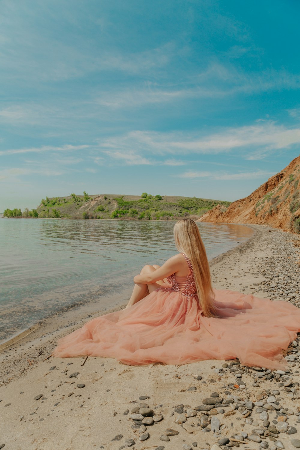 woman in pink dress sitting on brown sand near body of water during daytime