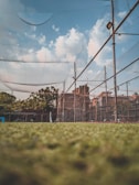 A sports net enclosure is surrounded by chain-link fencing under a blue sky with scattered clouds. The ground appears grassy, and some trees and buildings are visible in the background. Metal posts support the nets, and there is a tall light fixture on one of the poles.