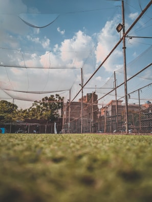 A sports net enclosure is surrounded by chain-link fencing under a blue sky with scattered clouds. The ground appears grassy, and some trees and buildings are visible in the background. Metal posts support the nets, and there is a tall light fixture on one of the poles.