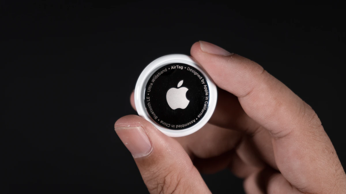 Hand holding an Apple AirTag against a dark background, showing the Apple logo and AirTag engraving