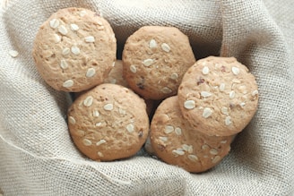 A group of oatmeal cookies with visible oats is placed on a textured burlap fabric. The cookies have a golden-brown color and are arranged closely together, creating a rustic and homemade appearance.