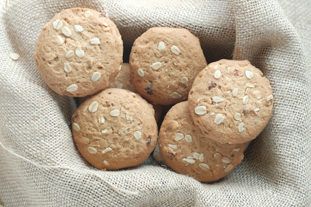 A group of oatmeal cookies with visible oats is placed on a textured burlap fabric. The cookies have a golden-brown color and are arranged closely together, creating a rustic and homemade appearance.