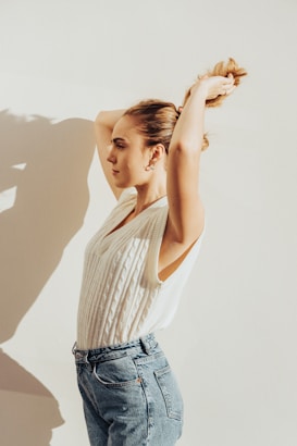 A woman with tied back hair is stretching her arms above her head against a plain light background. She wears a sleeveless white knit top paired with high-waisted blue jeans. The lighting creates a soft shadow behind her.