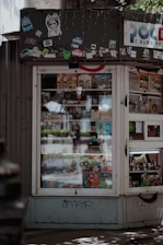 A small kiosk or newsstand with a window displaying various magazines and food items. The kiosk is adorned with multiple stickers and graffiti at the top, which gives it an urban, eclectic feel. Sunlight and shadows create a pattern on the ground in front of the kiosk.