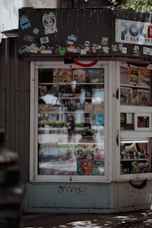 A small kiosk or newsstand with a window displaying various magazines and food items. The kiosk is adorned with multiple stickers and graffiti at the top, which gives it an urban, eclectic feel. Sunlight and shadows create a pattern on the ground in front of the kiosk.