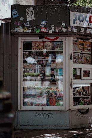 A small kiosk or newsstand with a window displaying various magazines and food items. The kiosk is adorned with multiple stickers and graffiti at the top, which gives it an urban, eclectic feel. Sunlight and shadows create a pattern on the ground in front of the kiosk.