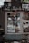 A small kiosk or newsstand with a window displaying various magazines and food items. The kiosk is adorned with multiple stickers and graffiti at the top, which gives it an urban, eclectic feel. Sunlight and shadows create a pattern on the ground in front of the kiosk.
