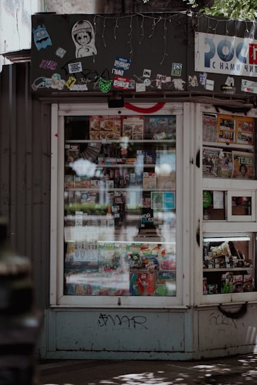 A small kiosk or newsstand with a window displaying various magazines and food items. The kiosk is adorned with multiple stickers and graffiti at the top, which gives it an urban, eclectic feel. Sunlight and shadows create a pattern on the ground in front of the kiosk.