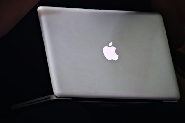 A sleek silver laptop with a prominent glowing Apple logo on its lid, placed on a dark surface.