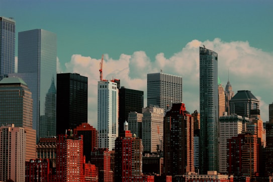 A vibrant city skyline with a mix of modern and older skyscrapers. The scene captures tall buildings with glass facades and brick structures under a partly cloudy sky. The architectural diversity is highlighted by various heights and designs.