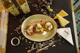 A table setting with an oval plate containing three pieces of food garnished with herbs and edible flowers. Wooden table partially covered with a black placemat, adorned with two bottles of olive oil, triangular wedges of cheese, a small bowl of peppercorns, and a pinecone. Also visible are decorative smiling fruits or gummies and a folded napkin with 'ALMA Bakery & Cafe' branding. The scene is completed with scattered salt crystals.