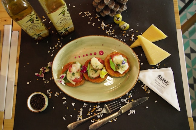 A table setting with an oval plate containing three pieces of food garnished with herbs and edible flowers. Wooden table partially covered with a black placemat, adorned with two bottles of olive oil, triangular wedges of cheese, a small bowl of peppercorns, and a pinecone. Also visible are decorative smiling fruits or gummies and a folded napkin with 'ALMA Bakery & Cafe' branding. The scene is completed with scattered salt crystals.