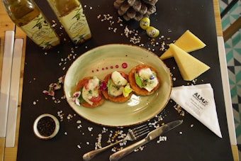 A table setting with an oval plate containing three pieces of food garnished with herbs and edible flowers. Wooden table partially covered with a black placemat, adorned with two bottles of olive oil, triangular wedges of cheese, a small bowl of peppercorns, and a pinecone. Also visible are decorative smiling fruits or gummies and a folded napkin with 'ALMA Bakery & Cafe' branding. The scene is completed with scattered salt crystals.
