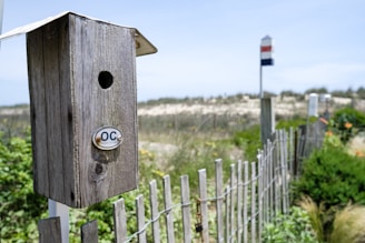 A weathered wooden birdhouse with a circular entrance hole mounted on a white pole. The birdhouse features a small metal plate with the letters 'OC' emblazoned on it. In the background, a sandy landscape with sparse vegetation and a wooden fence are visible, with another birdhouse, painted in red, white, and blue stripes, perched atop a distant pole.