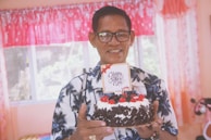 An elderly man wearing glasses and a shirt with a palm tree pattern is holding a chocolate cake decorated with cherries and cream. A small card on top of the cake reads 'Happy Father's Day'. The background shows bright curtains and a window.