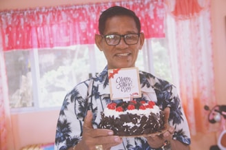 An elderly man wearing glasses and a shirt with a palm tree pattern is holding a chocolate cake decorated with cherries and cream. A small card on top of the cake reads 'Happy Father's Day'. The background shows bright curtains and a window.