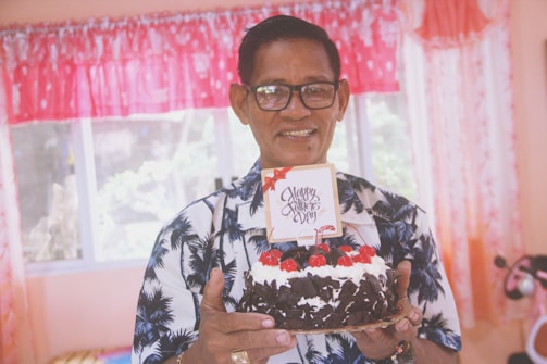 An elderly man wearing glasses and a shirt with a palm tree pattern is holding a chocolate cake decorated with cherries and cream. A small card on top of the cake reads 'Happy Father's Day'. The background shows bright curtains and a window.