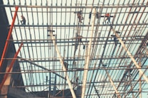 Workers are perched atop a complex network of scaffolding beams under a bright sky, engaging in construction or maintenance tasks.