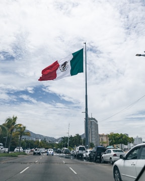 A large flag of Mexico is prominently displayed on a tall flagpole, fluttering in the breeze. The background features a bustling cityscape with numerous cars lining the road, palm trees on the sidewalk, and several tall buildings. The sky is mostly cloudy with patches of blue visible.