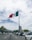 A large flag of Mexico is prominently displayed on a tall flagpole, fluttering in the breeze. The background features a bustling cityscape with numerous cars lining the road, palm trees on the sidewalk, and several tall buildings. The sky is mostly cloudy with patches of blue visible.