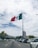 A large flag of Mexico is prominently displayed on a tall flagpole, fluttering in the breeze. The background features a bustling cityscape with numerous cars lining the road, palm trees on the sidewalk, and several tall buildings. The sky is mostly cloudy with patches of blue visible.