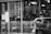 A black and white image showing a tattoo studio with large windows displaying tattoo designs. There is a mannequin or a person wearing a face mask and a face shield, sitting inside the shop. The interior includes hanging decorations and various objects related to tattooing. Signs on the window detail safety guidelines and restrictions.