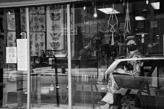 A black and white image showing a tattoo studio with large windows displaying tattoo designs. There is a mannequin or a person wearing a face mask and a face shield, sitting inside the shop. The intehttps://kcolestudio.com/wp-content/uploads/2024/06/woman-traveling-in-france-2023-11-27-05-16-47-utc_Easy-Resize.com_.jpgr includes hanging decorations and vahttps://kcolestudio.com/wp-content/uploads/2024/06/woman-traveling-in-france-2023-11-27-05-16-47-utc_Easy-Resize.com_.jpgus objects related to tattooing. Signs on the window detail safety guidelines and restrictions.