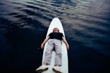 A person is lying on their back on a white stand-up paddleboard, floating on calm, dark water. The individual is wearing a black shirt, white pants, and white shoes. The surface of the water has gentle ripples, creating a tranquil atmosphere.