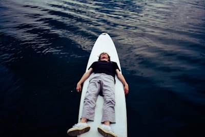 A person is lying on their back on a white stand-up paddleboard, floating on calm, dark water. The individual is wearing a black shirt, white pants, and white shoes. The surface of the water has gentle ripples, creating a tranquil atmosphere.