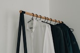 A minimalist clothing rack holding several hangers with garments, including a prominently visible white shirt and several dark-colored jackets. The rack is supported by a simple black metal frame and wooden rod, set against a plain white wall.