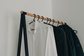 A minimalist clothing rack holding several hangers with garments, including a prominently visible white shirt and several dark-colored jackets. The rack is supported by a simple black metal frame and wooden rod, set against a plain white wall.