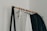 A minimalist clothing rack holding several hangers with garments, including a prominently visible white shirt and several dark-colored jackets. The rack is supported by a simple black metal frame and wooden rod, set against a plain white wall.
