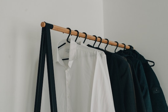 A minimalist clothing rack holding several hangers with garments, including a prominently visible white shirt and several dark-colored jackets. The rack is supported by a simple black metal frame and wooden rod, set against a plain white wall.