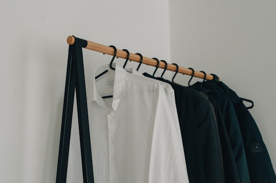 A minimalist clothing rack holding several hangers with garments, including a prominently visible white shirt and several dark-colored jackets. The rack is supported by a simple black metal frame and wooden rod, set against a plain white wall.