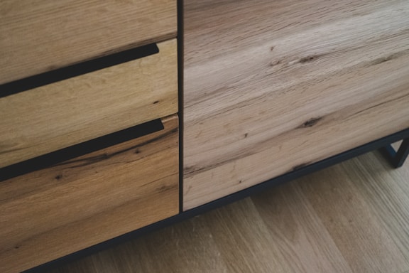 A close-up view of wooden furniture showcasing two drawers with integrated black handles. The surface texture of the wood is smooth with visible natural grain patterns and knots. The color tone is a mixture of light and medium brown, suggesting natural wood.