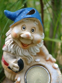 A cheerful garden gnome figurine with a blue pointed hat and a white beard. The gnome is holding a round object, possibly a light, and is set against a backdrop of green plants.