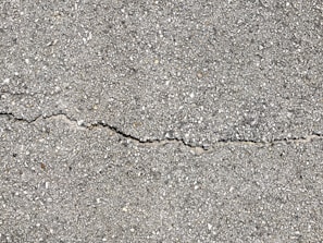 A close-up view of a textured gray concrete surface with a noticeable crack running horizontally across the image. The surface is rough with small pebbles and aggregates visible within the concrete material.