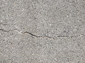 A close-up view of a textured gray concrete surface with a noticeable crack running horizontally across the image. The surface is rough with small pebbles and aggregates visible within the concrete material.
