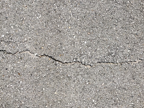 A close-up view of a textured gray concrete surface with a noticeable crack running horizontally across the image. The surface is rough with small pebbles and aggregates visible within the concrete material.
