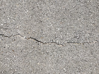 A close-up view of a textured gray concrete surface with a noticeable crack running horizontally across the image. The surface is rough with small pebbles and aggregates visible within the concrete material.